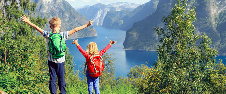 Ein Junge und ein Mädchen, von hinten zu sehen, strecken bei schönem Wetter die Arme auseinander; grüne Natur, ruhiges Gewässer und Berge vor ihnen.